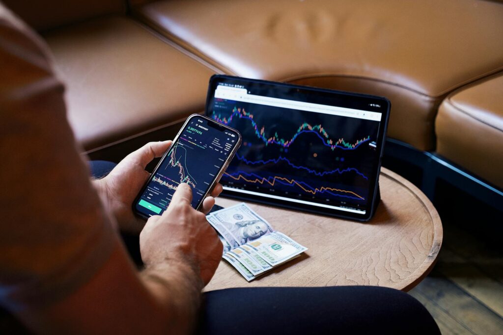 A person using a smartphone and tablet for stock market analysis, with graphs and cash visible.