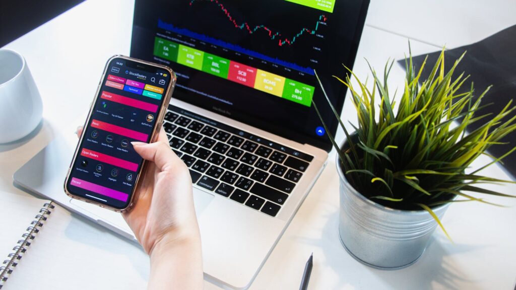 A person using a smartphone app to trade stocks with a laptop displaying market data. PER