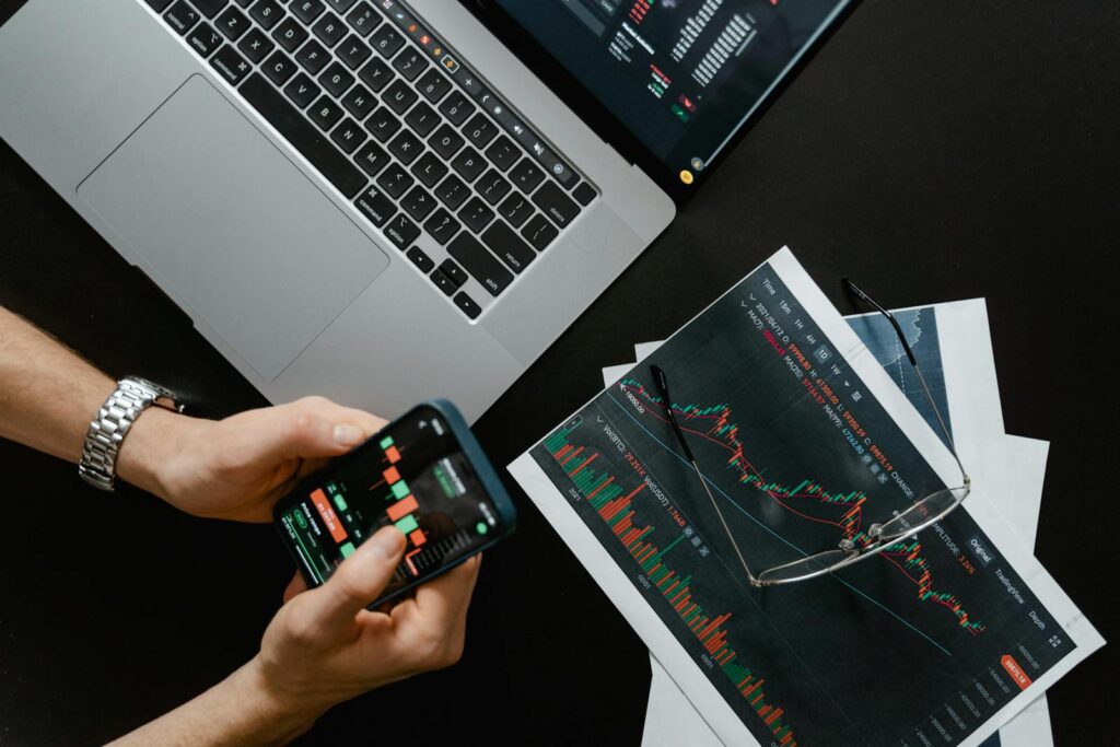 Hands using smartphone beside laptop with stock charts, showcasing digital trading. PER