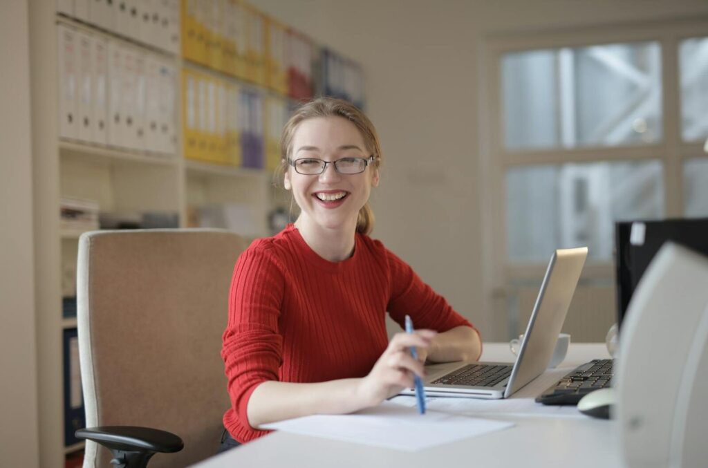 Happy woman in red sweater working at office desk with laptop and pen, smiling at camera, Comment épargner chaque mois